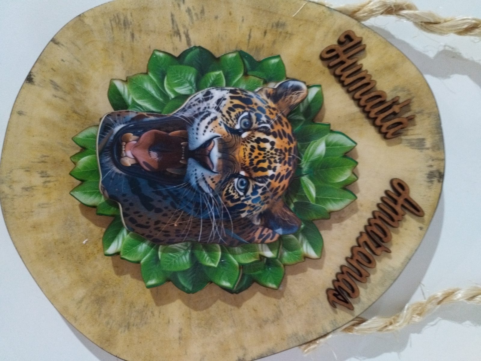 Placa decorativa de madeira com detalhe da onça-pintada em relevo e folhagem verde