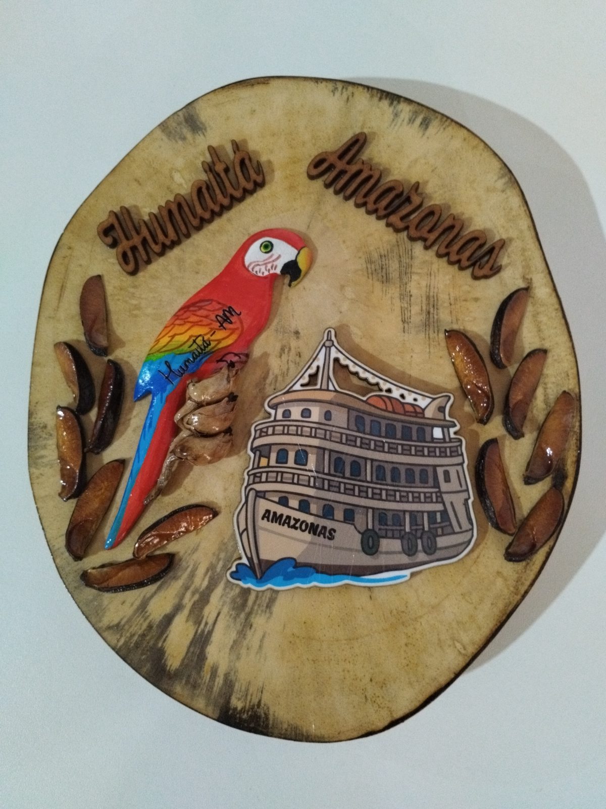 Plaquinha de madeira com arara-vermelha, barco, sementes e a frase Humaitá Amazonas
