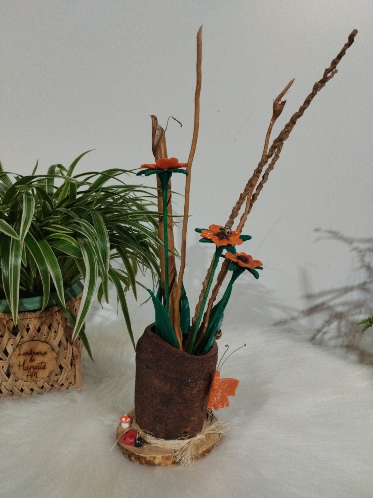 Vaso rústico visto de lado com três flores laranjas, folhas verdes e borboleta