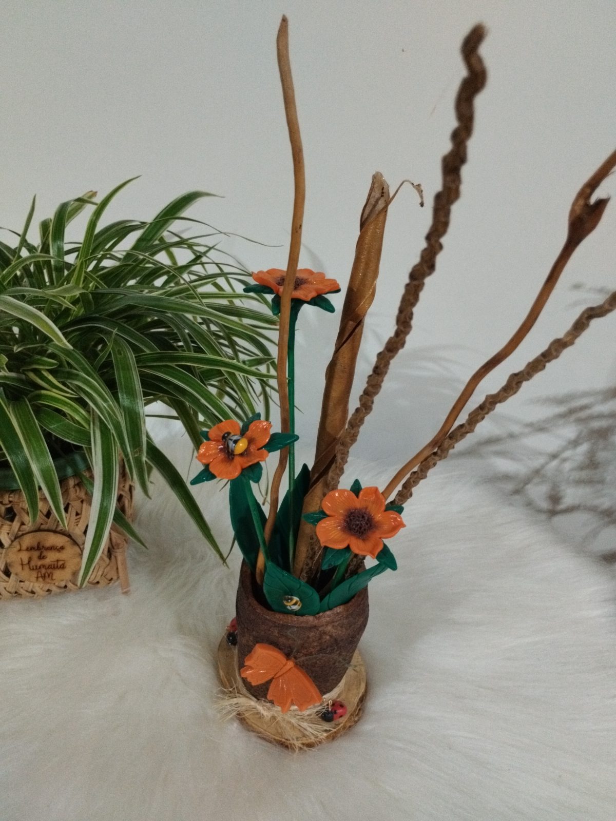 Vaso rústico com flores laranjas, folhas verdes, galhos secos e pequenos insetos decorativos