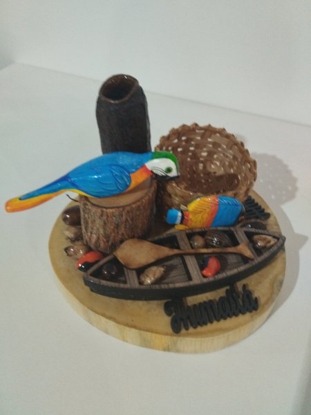 Enfeite de mesa com arara azul, canoa, cestinha e sementes sobre base de madeira.