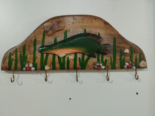 Porta-chaves de madeira em formato abaulado com desenho de pirarucu, vegetação verde, sementes e ganchos metálicos