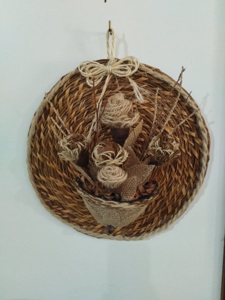 Enfeite de parede redondo feito de cipó ambé com flores de juta e laço de sisal ao centro.
