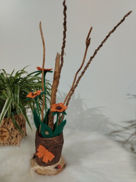 Vaso rústico marrom com flores laranjas, galhos secos e uma borboleta laranja aplicada na frente.