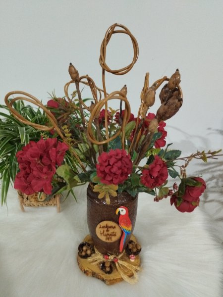 Vaso marrom rústico com flores vermelhas, galhos secos, uma arara e plaquinha escrita Lembrança de Humaitá AM.